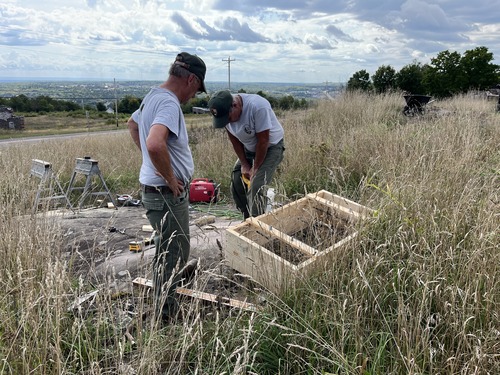 Two men stand in tall grass building a wood frame box.