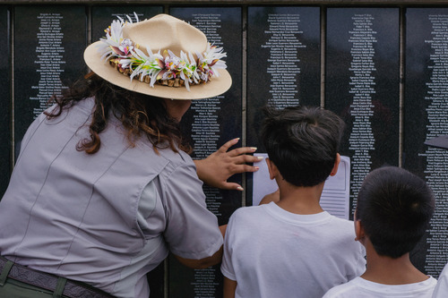 A woman in a park ranger uniform with flower around the brim of her hat helps two boys make a rubbing of a name on a black plaque covered with engraved names.