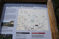 A blue metal panel with a white square on the upper left corner sits in a wooden frame. Words and images on the metal square give information about Copper Creek Trailhead. 