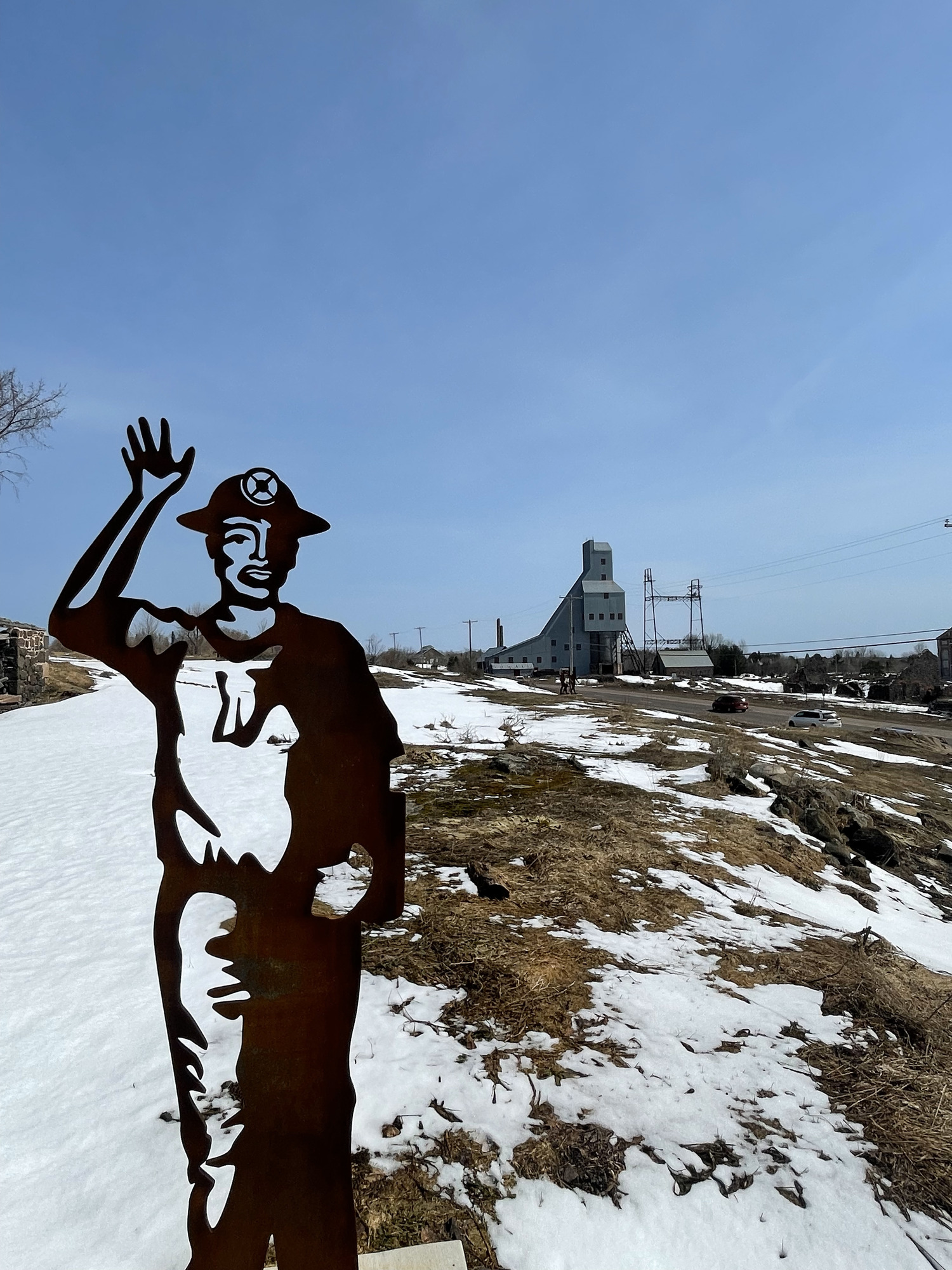 A sculpture of a miner waving in the foreground with the ruins of a mining buildings in the background. Snow covers the ground.