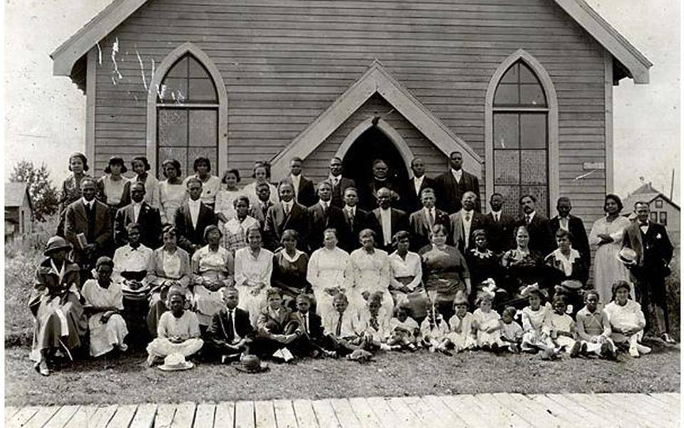 A black and white group photo in front of a simple, wooden church. The group shows men, women, and children posed in four rows. The children make up the first row and are seated in front of the adults.