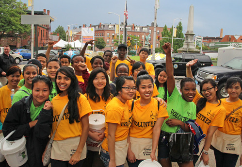 A group of young people pose, with some having their hands held high from peace signs to fist bumps. Their shirts are either yellow or green which has the Folk Festival label and "VOLUNTEER" read on it. Many are wearing small work aprons and are carrying donation buckets.