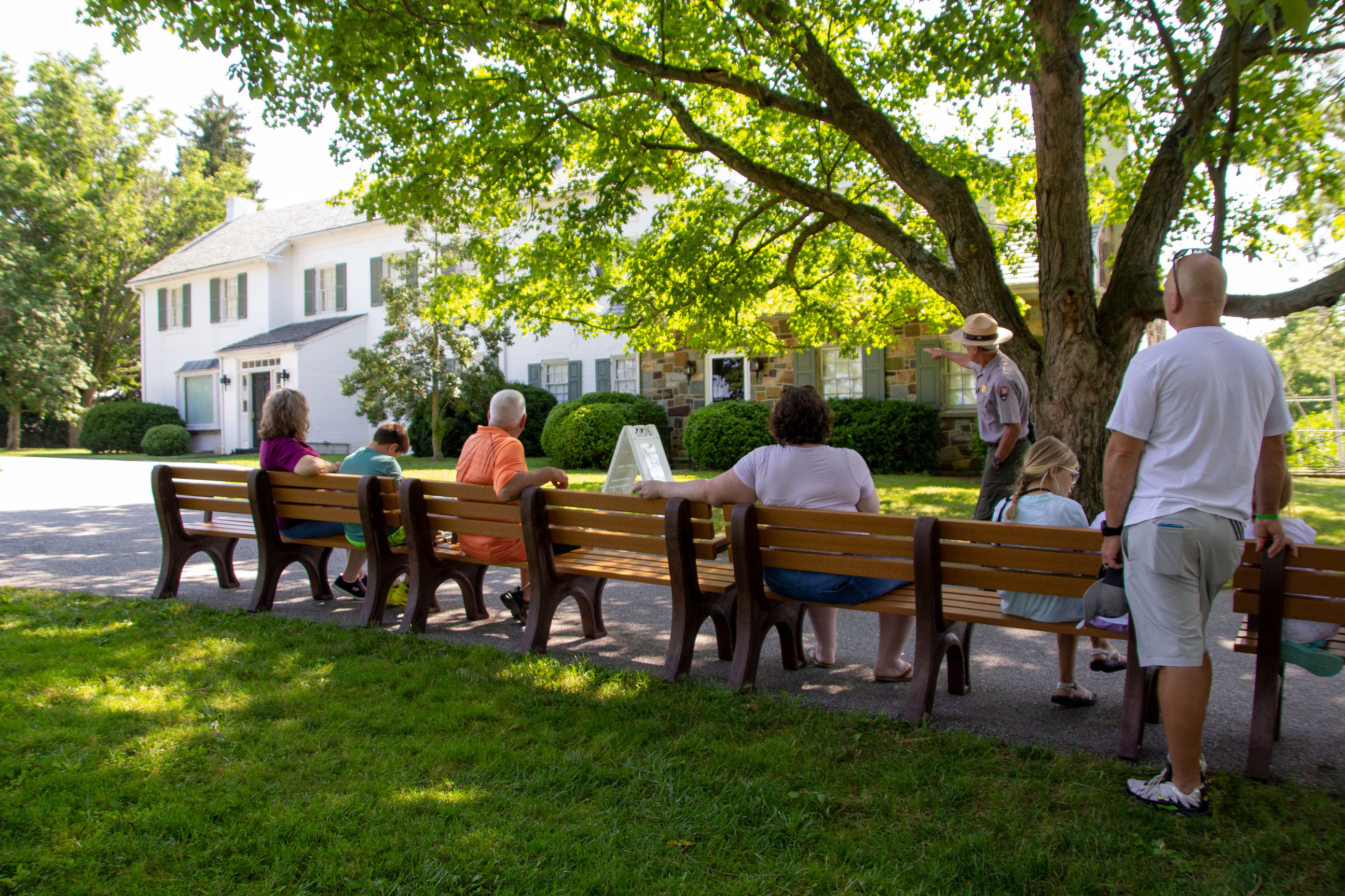 10 visitors sit on benches an watch a Park Ranger give a talk outside under shaded tree in front of a large white house.