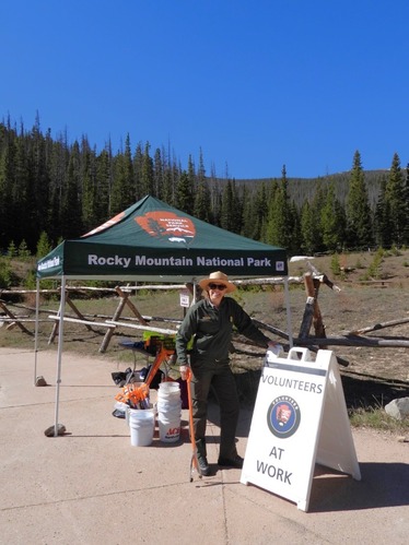 Park Ranger woman poses by a sign for a volunteer event outdoors with a blue sky, NPS pop up tent, and trees behind her. 
