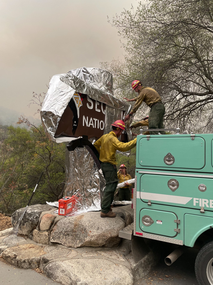 Three Firefighters wrap historic entrance sign for Sequoia National Park with silver fire shelter material to protect it from a wildfire. Oak trees and dense smoke from wildfire can be seen in the background.