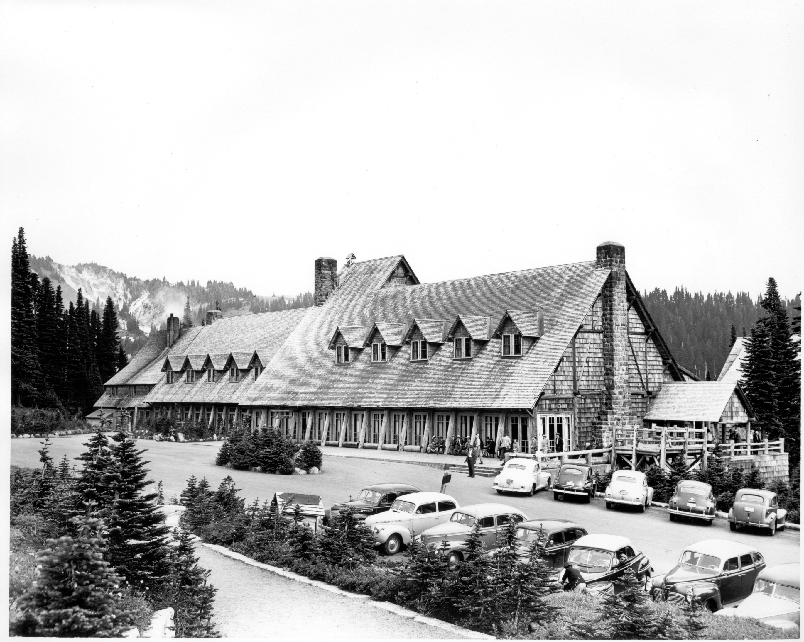 Historic photo. Rows of 1940s era cars parked in front a large building with an angled roof. 