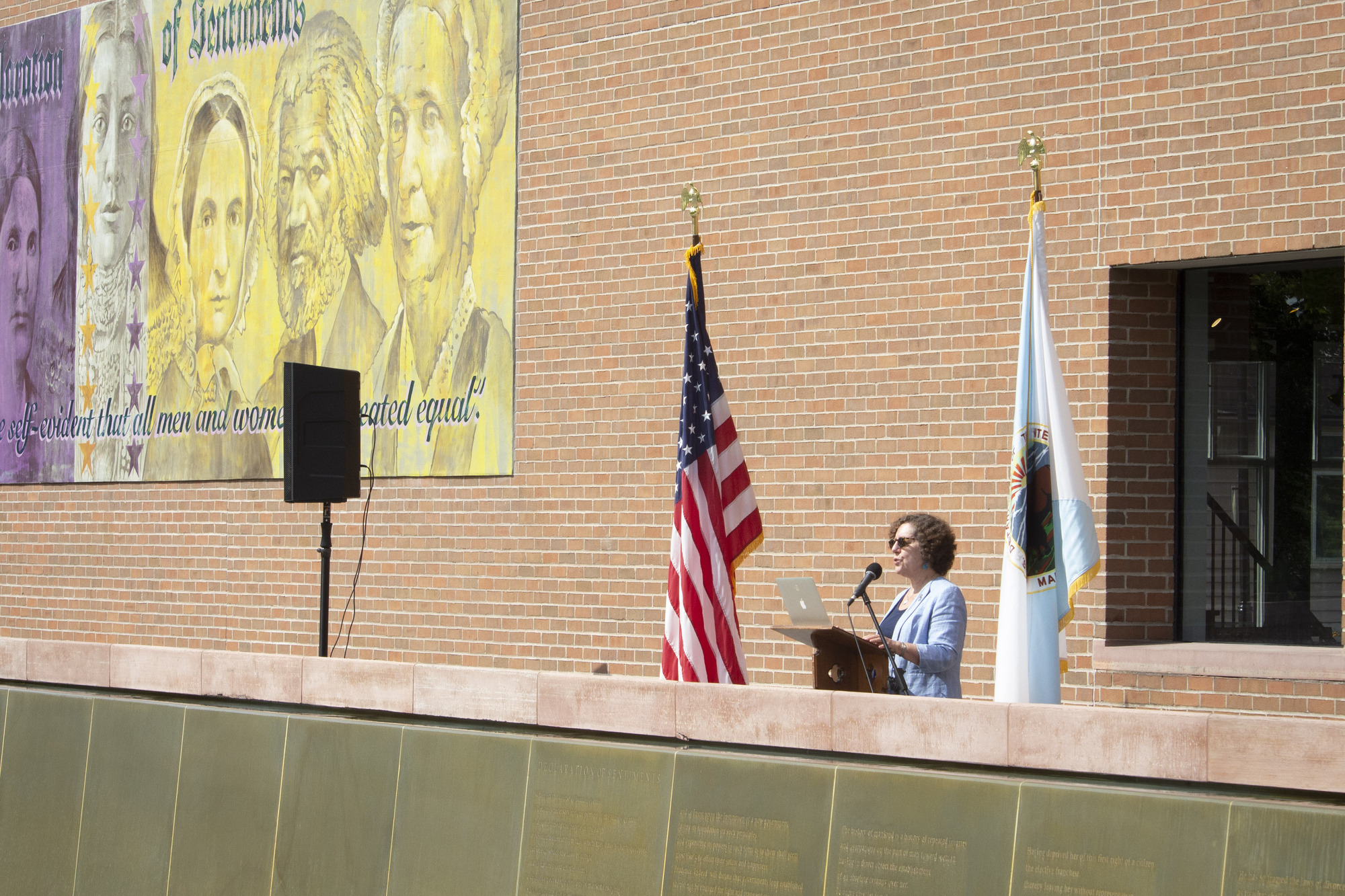 A woman stands at a lectern between two flags, in front of a brick wall.