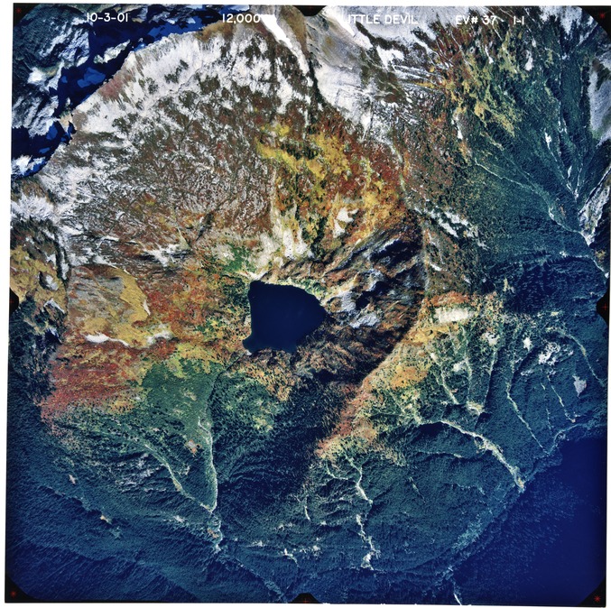 Color aerial photograph of glaciated mountain peak with alpine lake, forested valleys below.