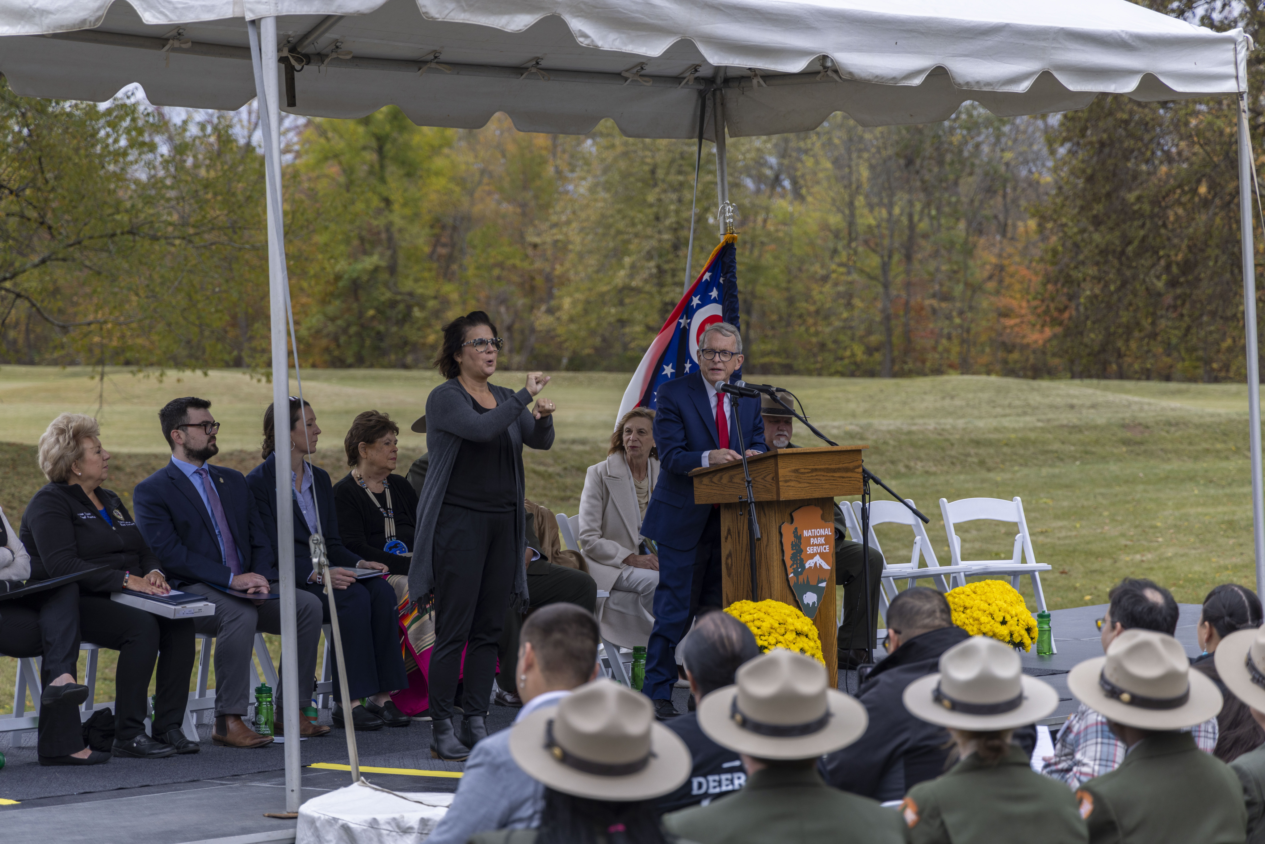 A lman in a blue suit stands at a wooden podium with the NPS arrowhead logo on the front as he speaks in to a microphone. Several people wearing tan flat hats can be seen sitting in front of the speaker at the podium.