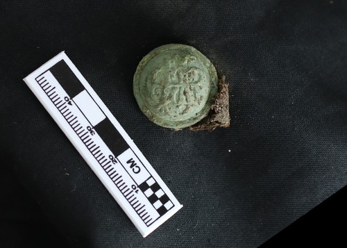 A round green button with brown highlights. There are raised letters, GAR, intertwined on it. On a black background with a ruler showing it as 3 cm in size.