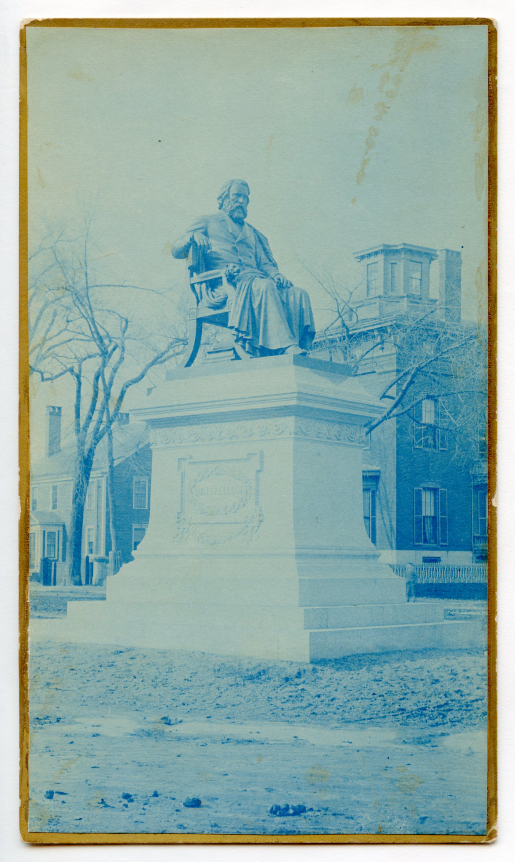 Blue-scale photograph of a life-size or larger statue of Henry Wadsworth Longfellow. Statue is on a tall pedestal outdoors. There are brick buildings in the background. Longfellow wears a suit and is draped in some kind of cloth, possibly a professor/graduation gown. He has chin-length hair and a full beard.