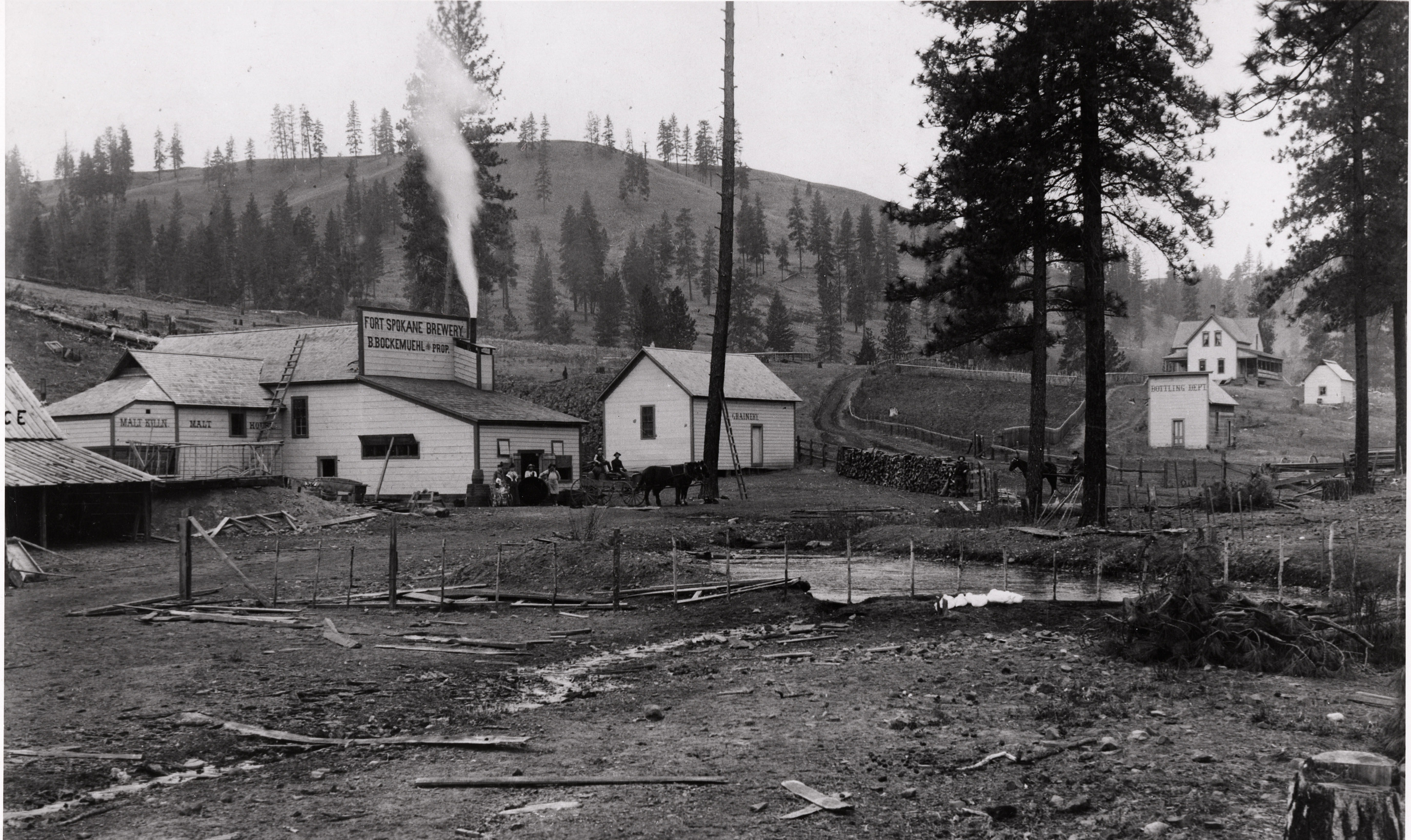 Black and white photograph of several plain wooden buildings clustered on a hill along a dirt road