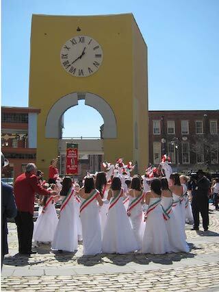 a small group of women, all dressed in floor length white dresses with red, green, and white sashes,  stand with their backs turned near a Italianate arch