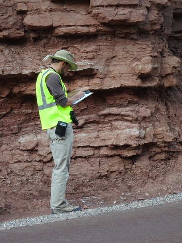 A young man in a yellow safety vest stands on the road side evaluating the red rock outcrop behind him. 