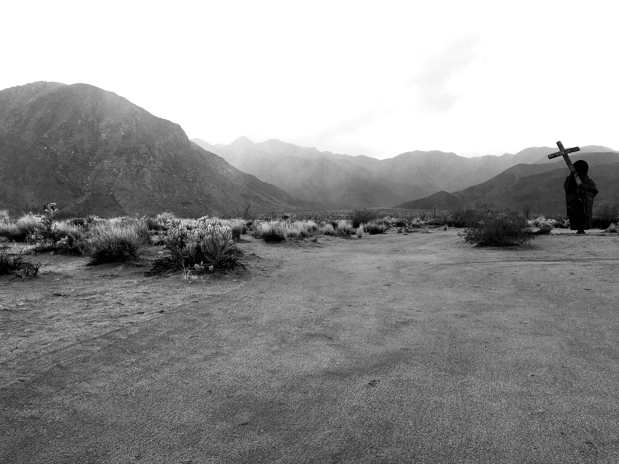 A black and white photo of a sculpture of a priest carrying a rosary and a cross in the desert