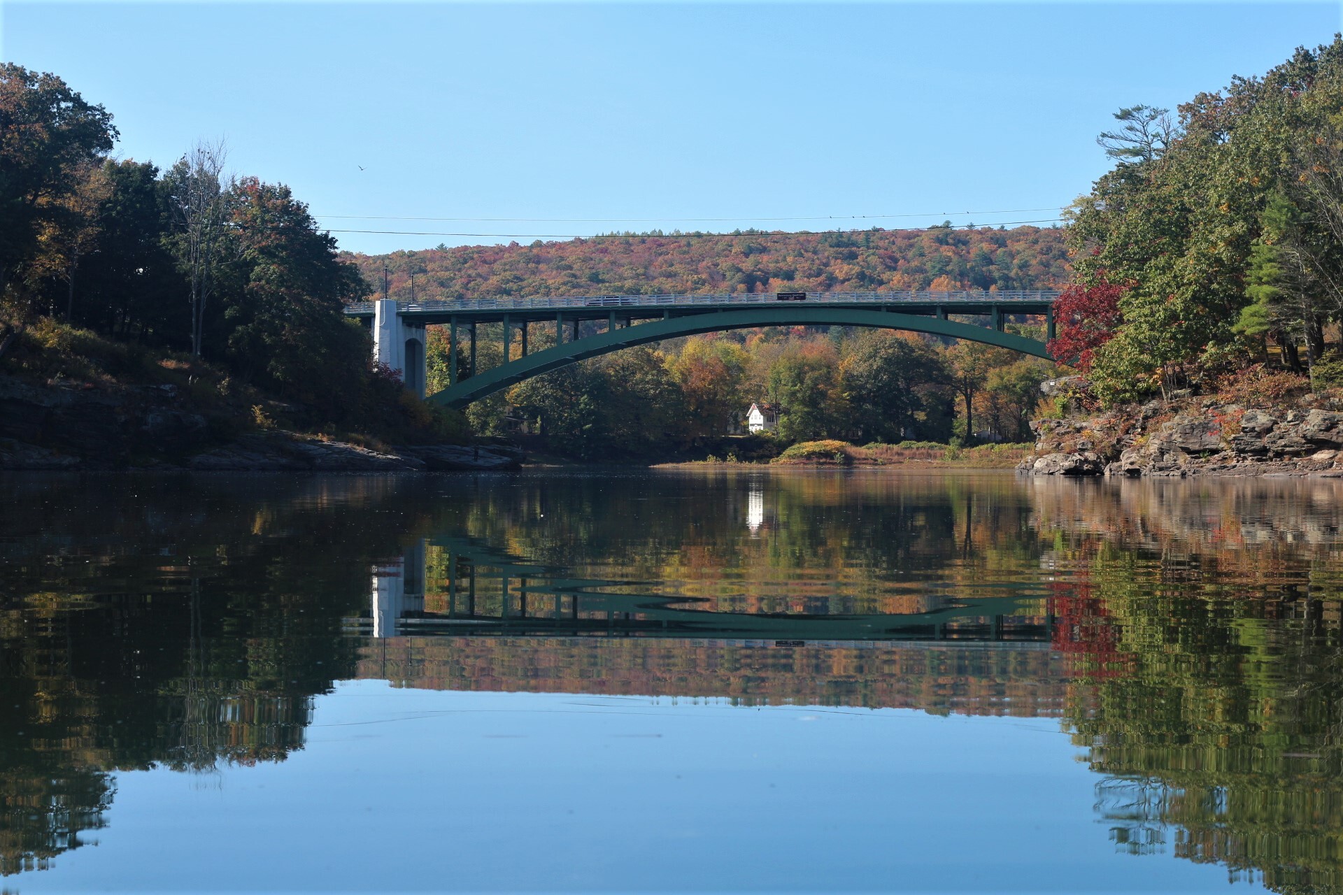 Flat vehicular bridge supported by green metal arch spans over river. In background is rolling forested hill colored orange, green, and red with leaves of autumn. The river reflects the bright blue sky and colors of fall.