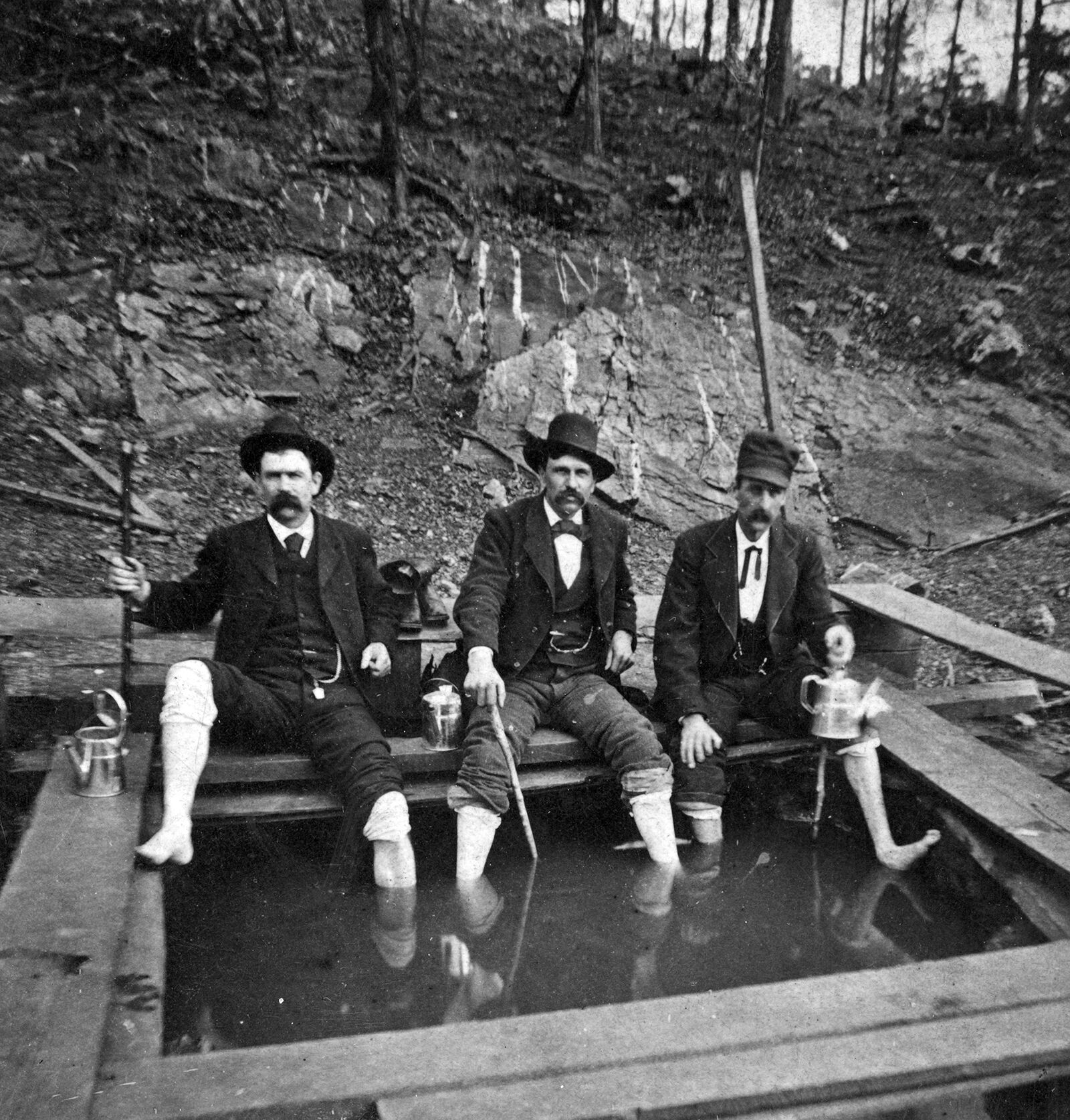 Three moustached men sit on a makeshift platform at the edge of a hot spring pool. They are wearing suits and have their pants rolled up so they can soak their feet in the water.