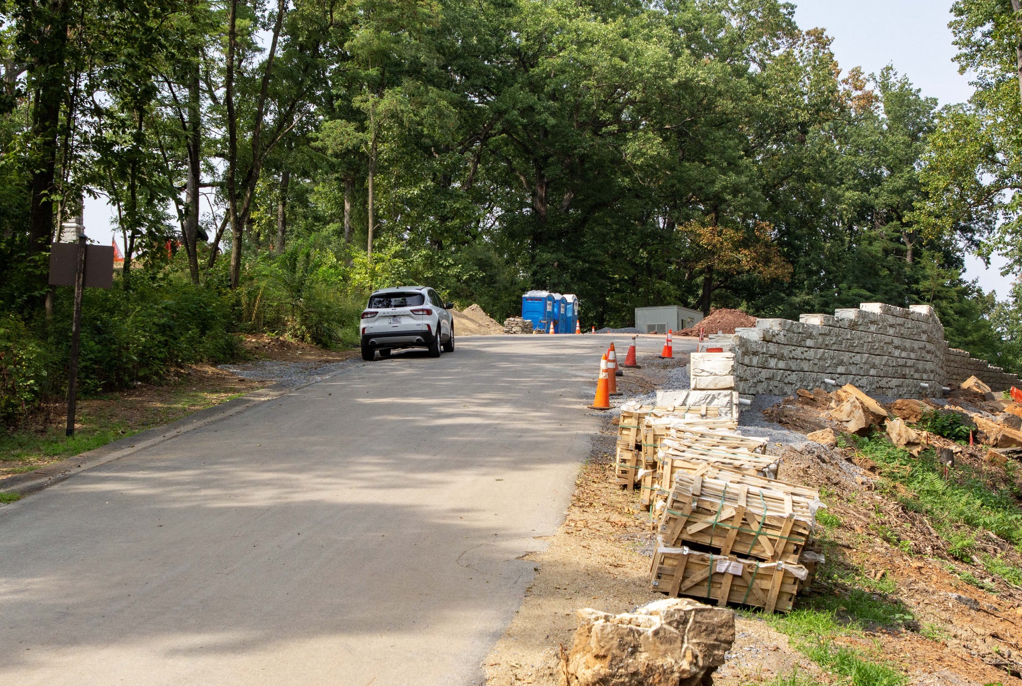 A white concrete retaining wall winds away from the camera along the right. A paved road runs through the center next to the retaining wall. A white, four-door car and three blue portable toilets are along the road on the left. Green trees surround the area.