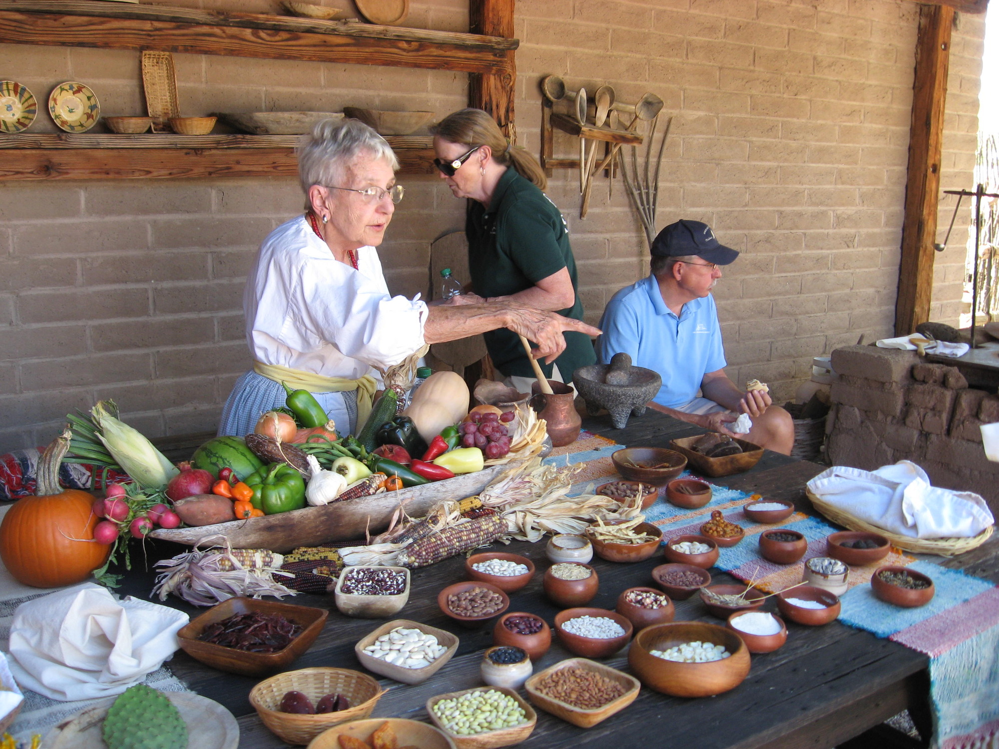 A person with short gray hair and glasses wearing a dress points from behind a table laid out with different types of beans, nuts, and vegetables