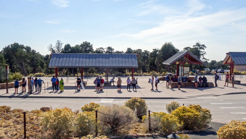 A large bus is pulling up to a bus stop with a line of people waiting to board. 