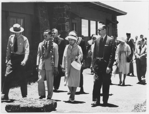 Black and white. A group of people walking in front of a building, accompanied by a park ranger