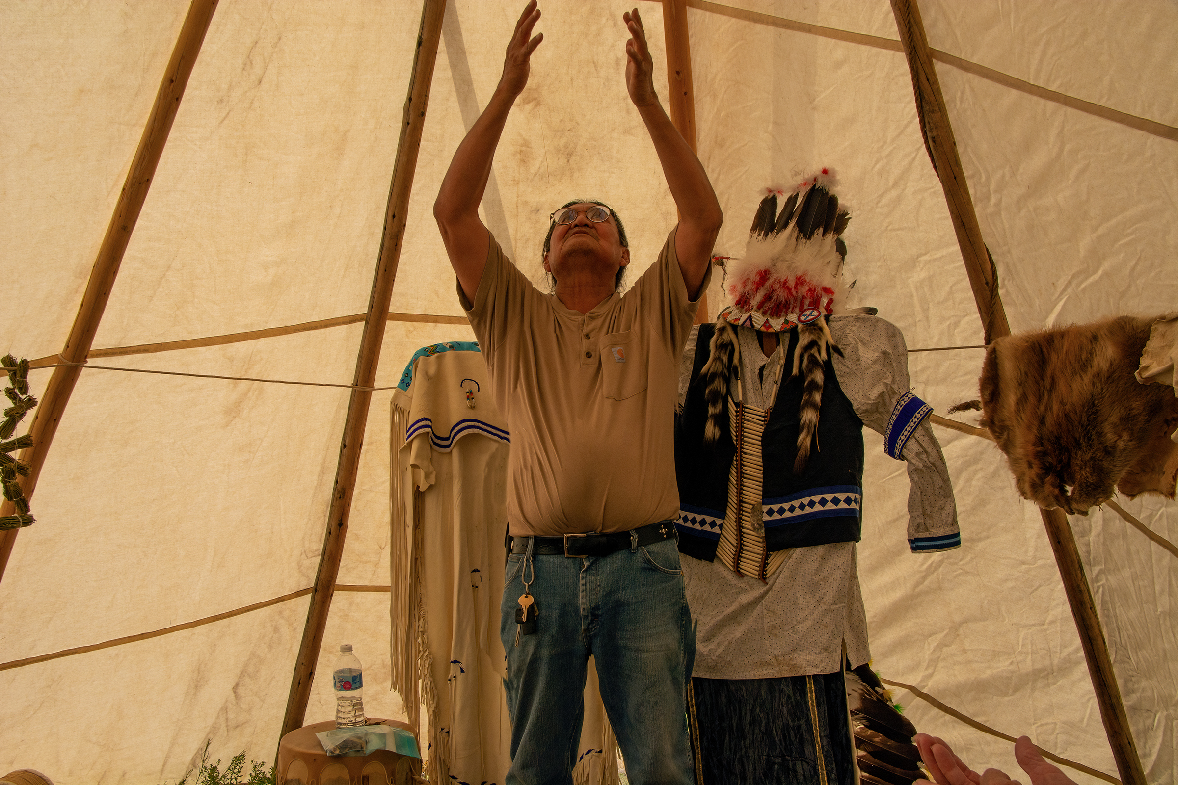 A Jicarilla Apache Elder lifts his hands while telling a traditional story within a tipi.  Behind him are traditional clothing items. 