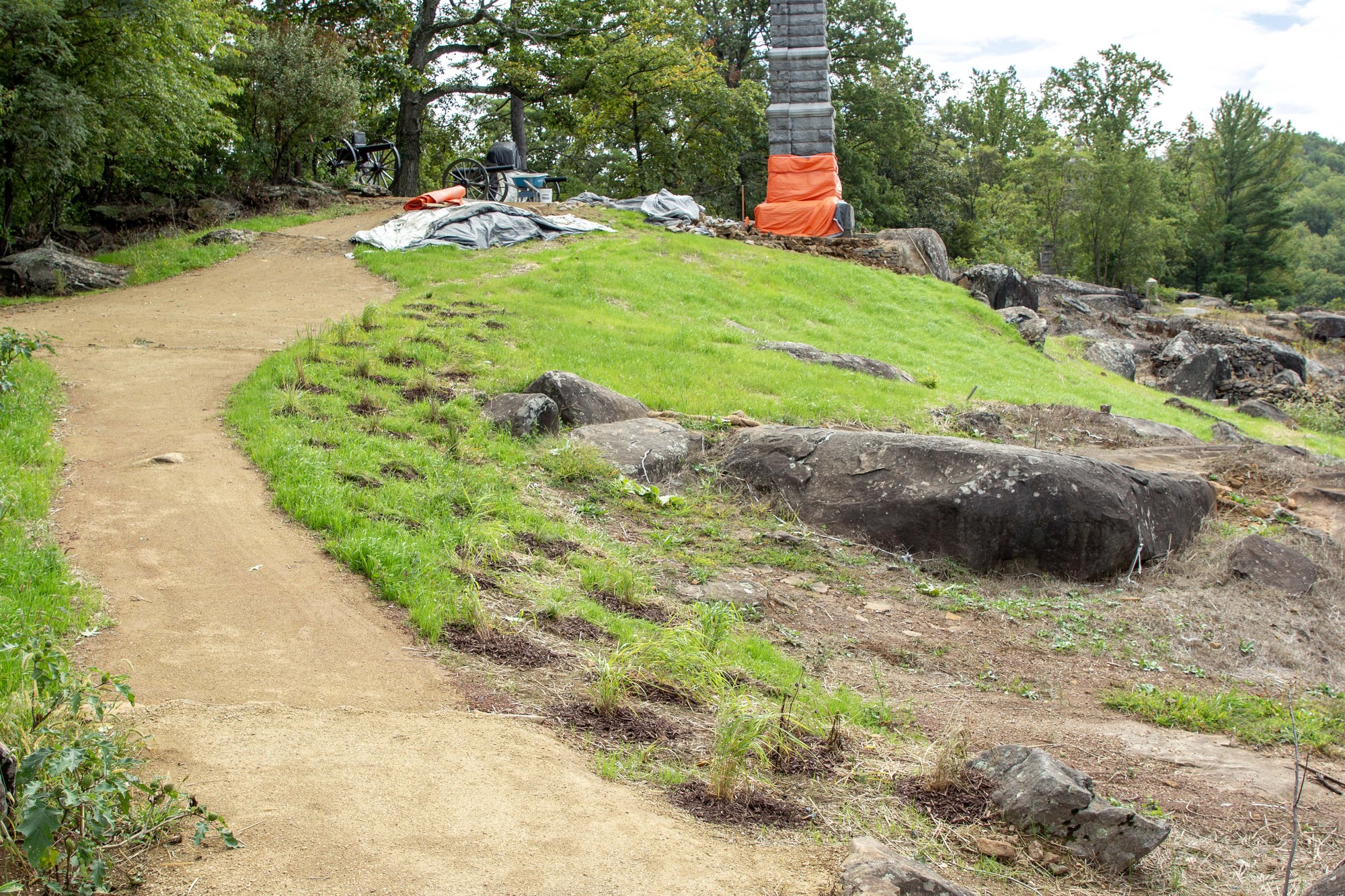 Lush green grass lines the slope of a hill between large boulders in the foreground and along the right of the photograph. Portions of two Civil War era cannons and a tall grey stone monument with a bright orange tarp wrapped around the bottom of the structure line the crest of the hill.