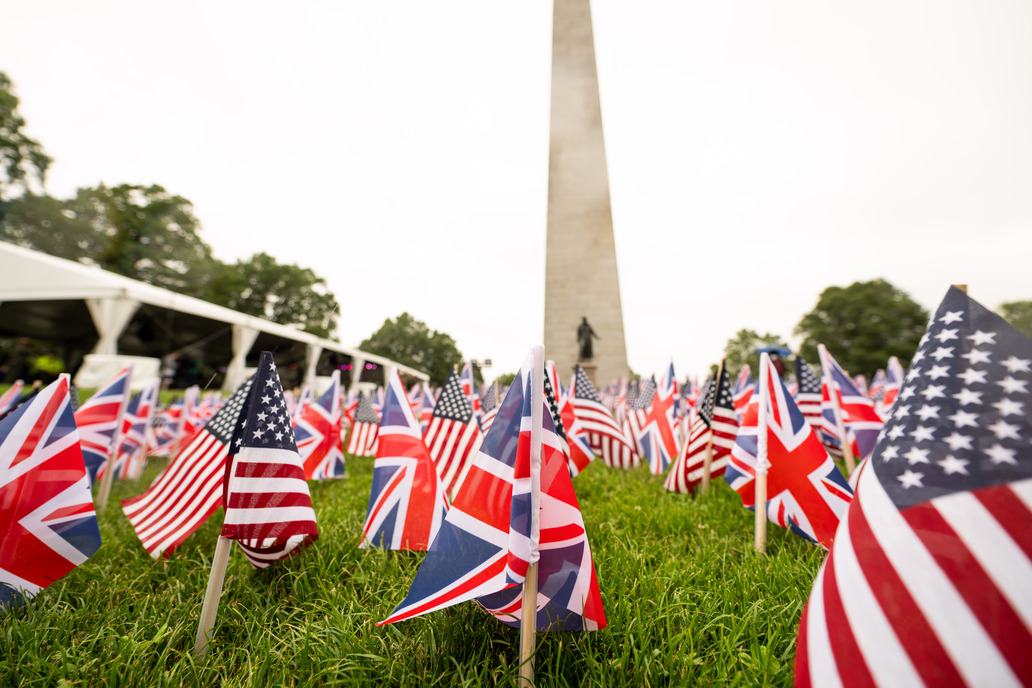 Rows of small US and UK Flags placed in a lawn. the Bunker Hill Monument is in the background.  