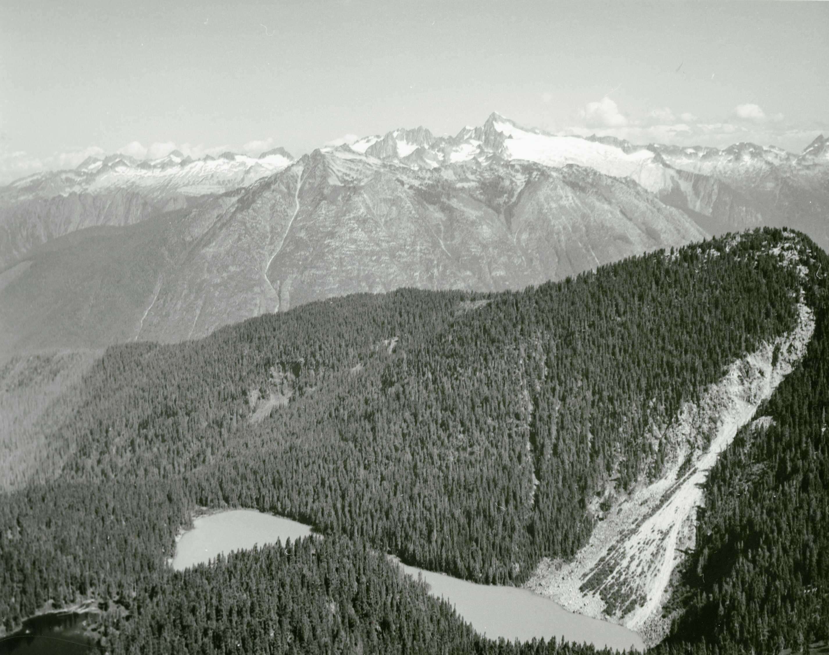 Two small lakes on the top of a forested mountain. Snow covered peaks in the background.