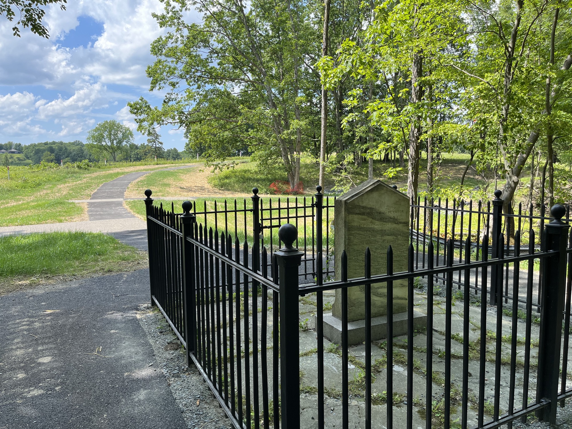 A stone monument surrounded by a black, iron fence dominates the foreground. Trees an a path leading into the distance are behind.