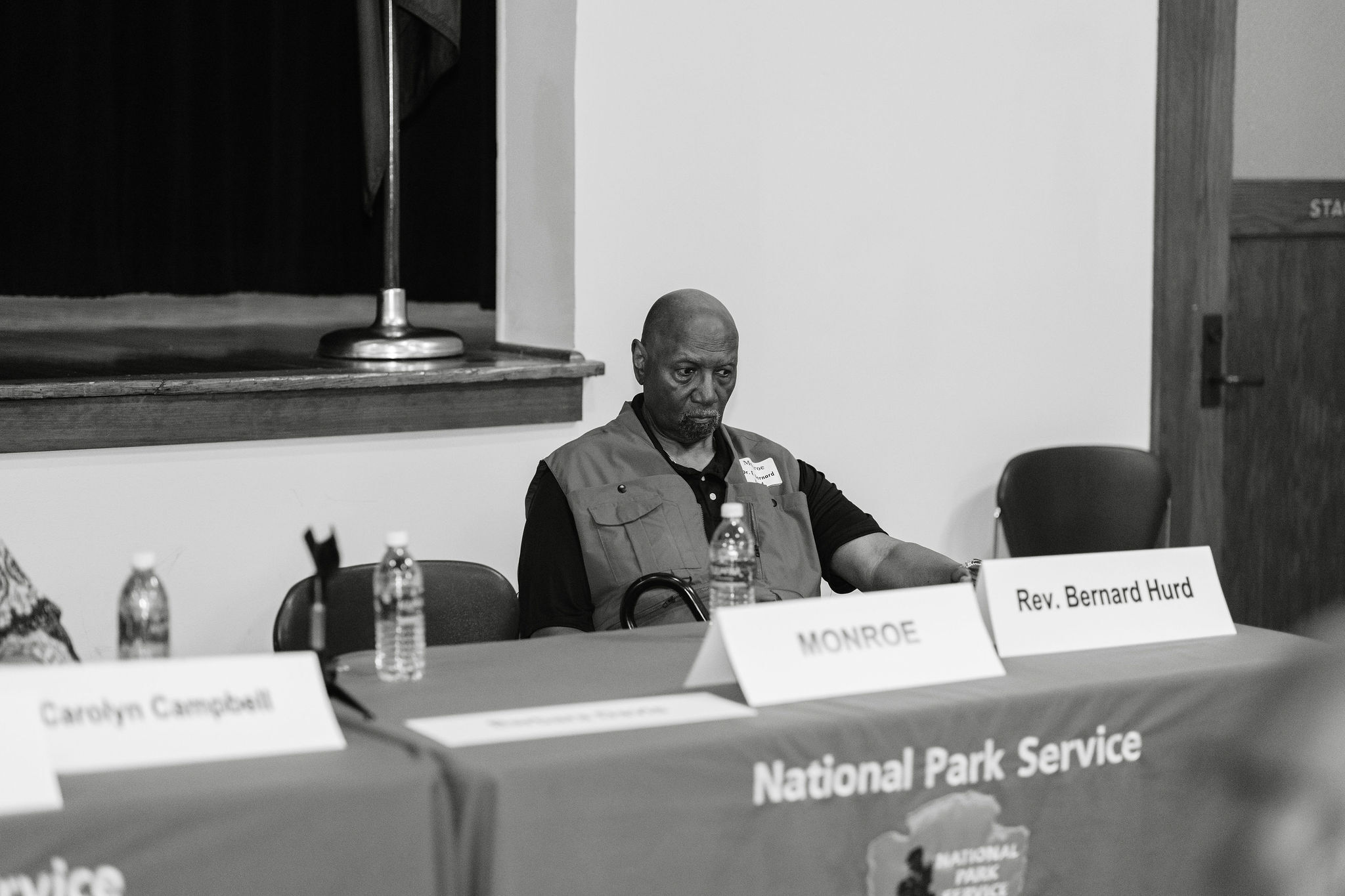Black and white photo of a man in a vest sitting at a table behind two table tents reading monroe on one and Rev. Bernard Hurd on another.
