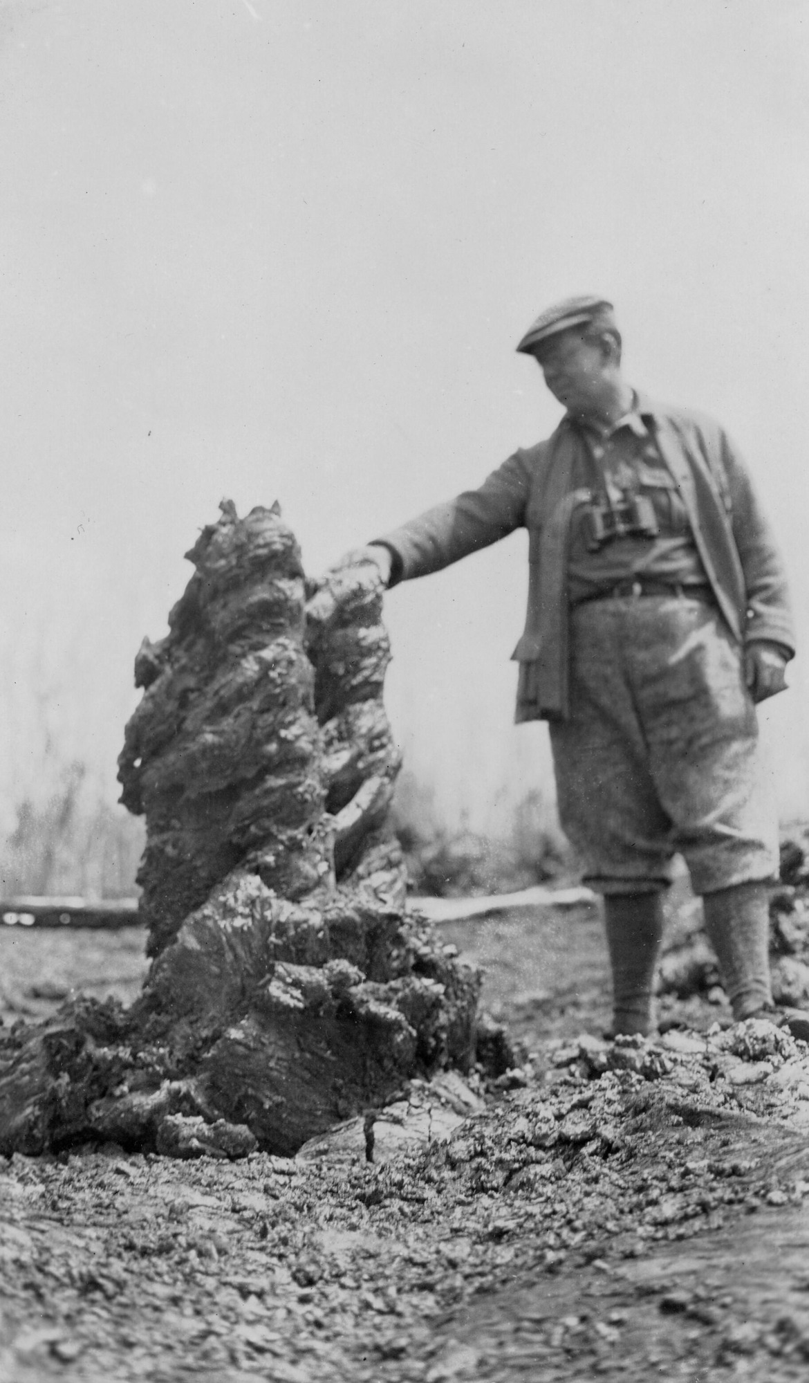 Black and white photograph of an unidentified man next to a tall lava formation. The man is on the right side of the image with binoculars around his neck, and his right hand is outstretched touching the top of the lava formation.  The lava formation extends vertically and emulates a thick tree trunk. The surrounding land is dry and rocky.