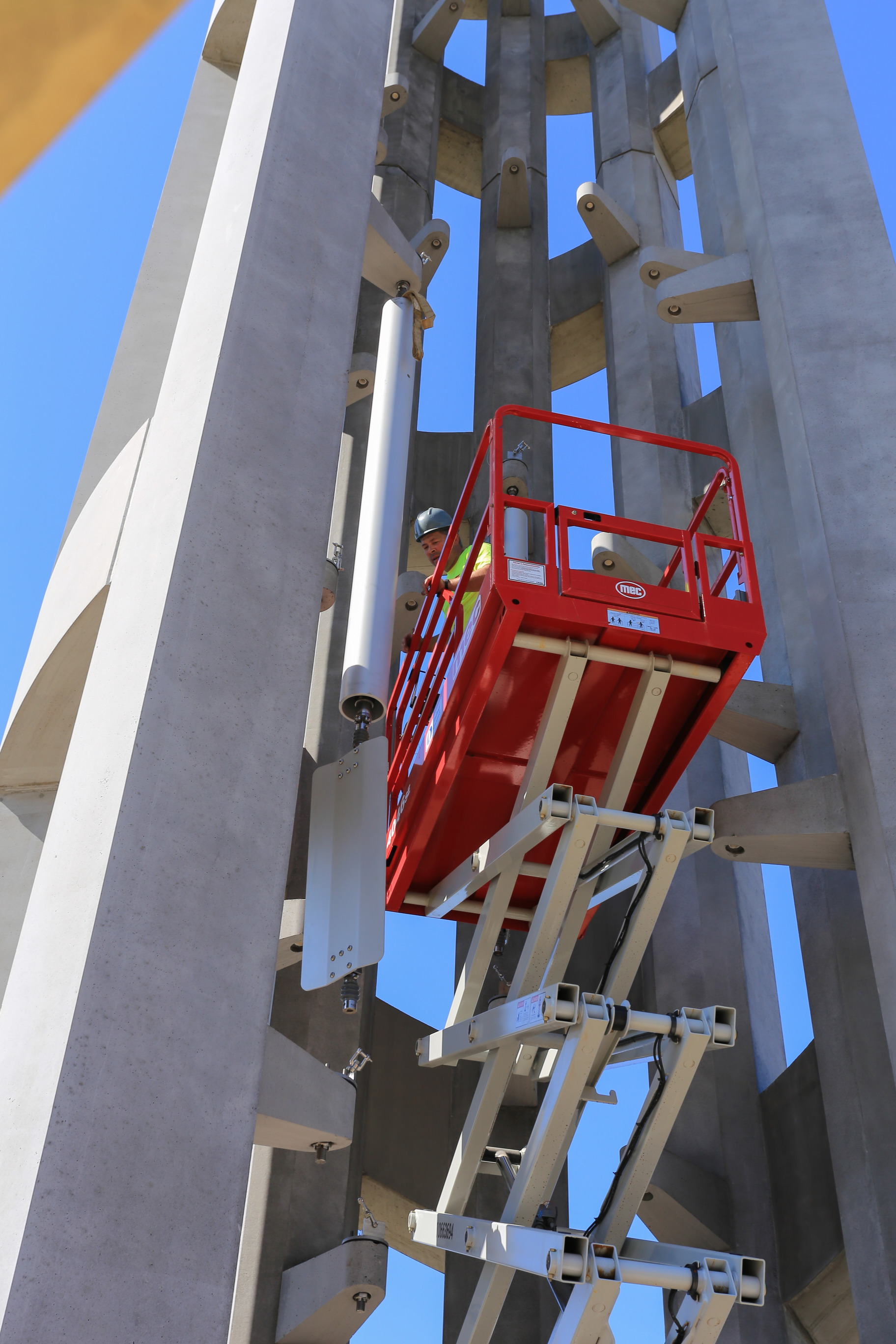 Man in hard hat on a red lift examines a long metal chime tube hanging in tall tower with many windows.