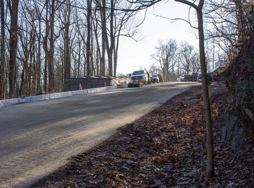 Large slabs of granite are aligned near the side of a roadway. A large stone wall appears near the line of granite. Vehicles are parked in a parking area in the upper right side of the photograph. 
