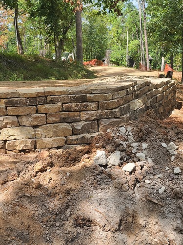 A stone retaining wall crosses the middle of the frame from left to right, then angles away at roughly a 60 degree angle from the center of the frame almost to the right edge in the distance. Below the wall in the foreground is bare earth and stone debris, and above the wall is a dirt road bed curving away into the distance with forest on either side and a cabin with a stone chimney just visible in the distance.