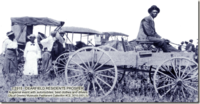 Black and white photo.  Man sits in open air wagon in suit and brimmed hat.  Other carriages, motorized, are lined in a row.  Women and men in the photo are all dressed in their Sunday best.  Word on photo read: c 1915 - Dearfield residents prosper, A special event with automobiles, best clothes and drivers. City of Greeley Museums Permanent Collection #C2_3015.0001.1c
