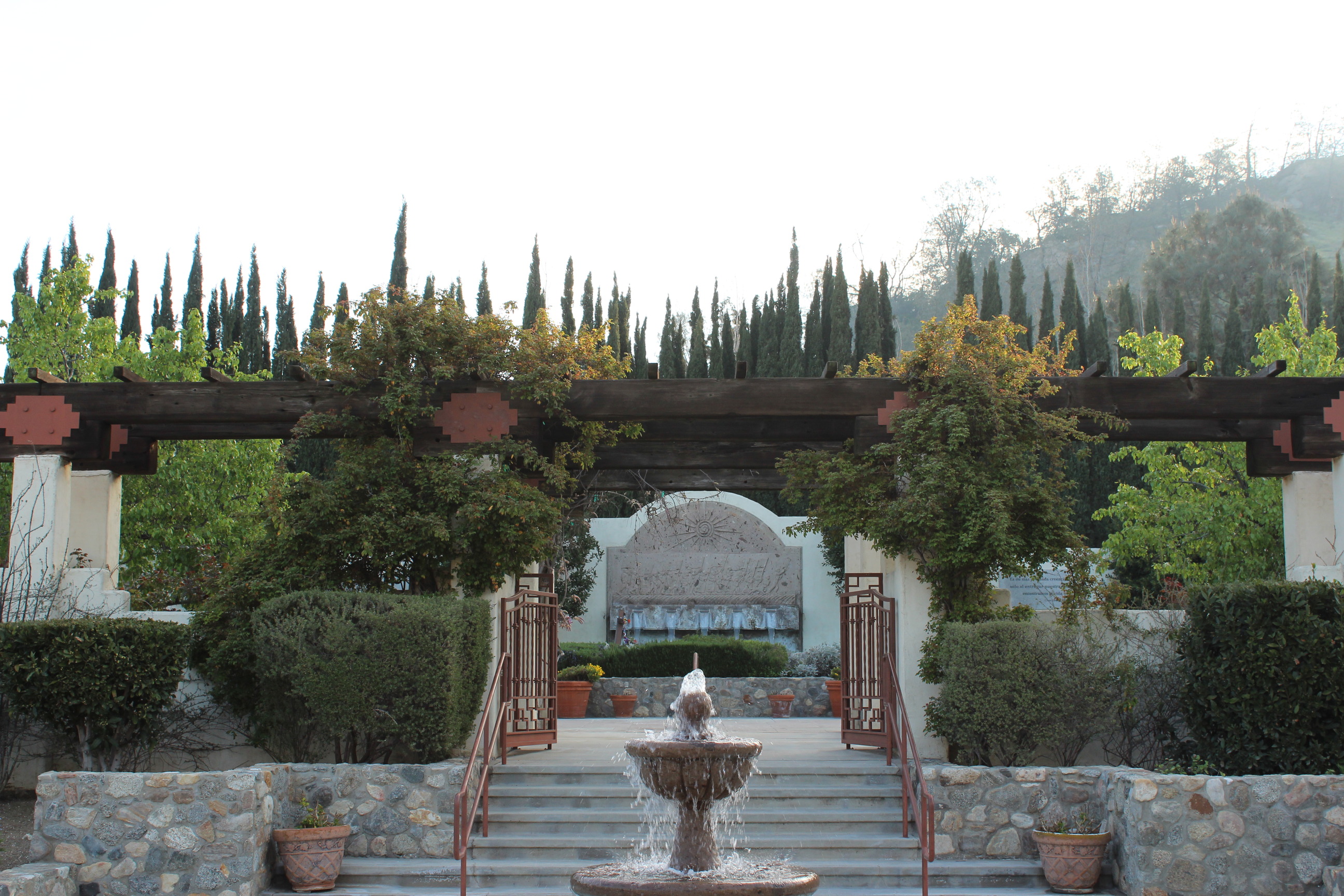 A fountain flows in front of a set of stairs that lead to a path that passes below a wooden arbor. In the background is a rectangular fountain with an arched top.