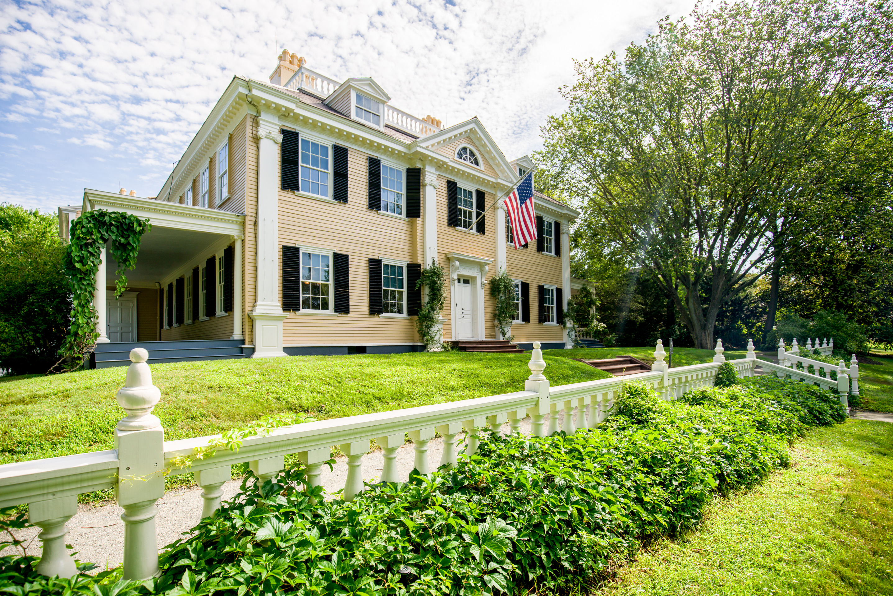 Yellow three-story mansion with symmetrical facade. White railing with green ivy in foreground, large elm tree to right of house.