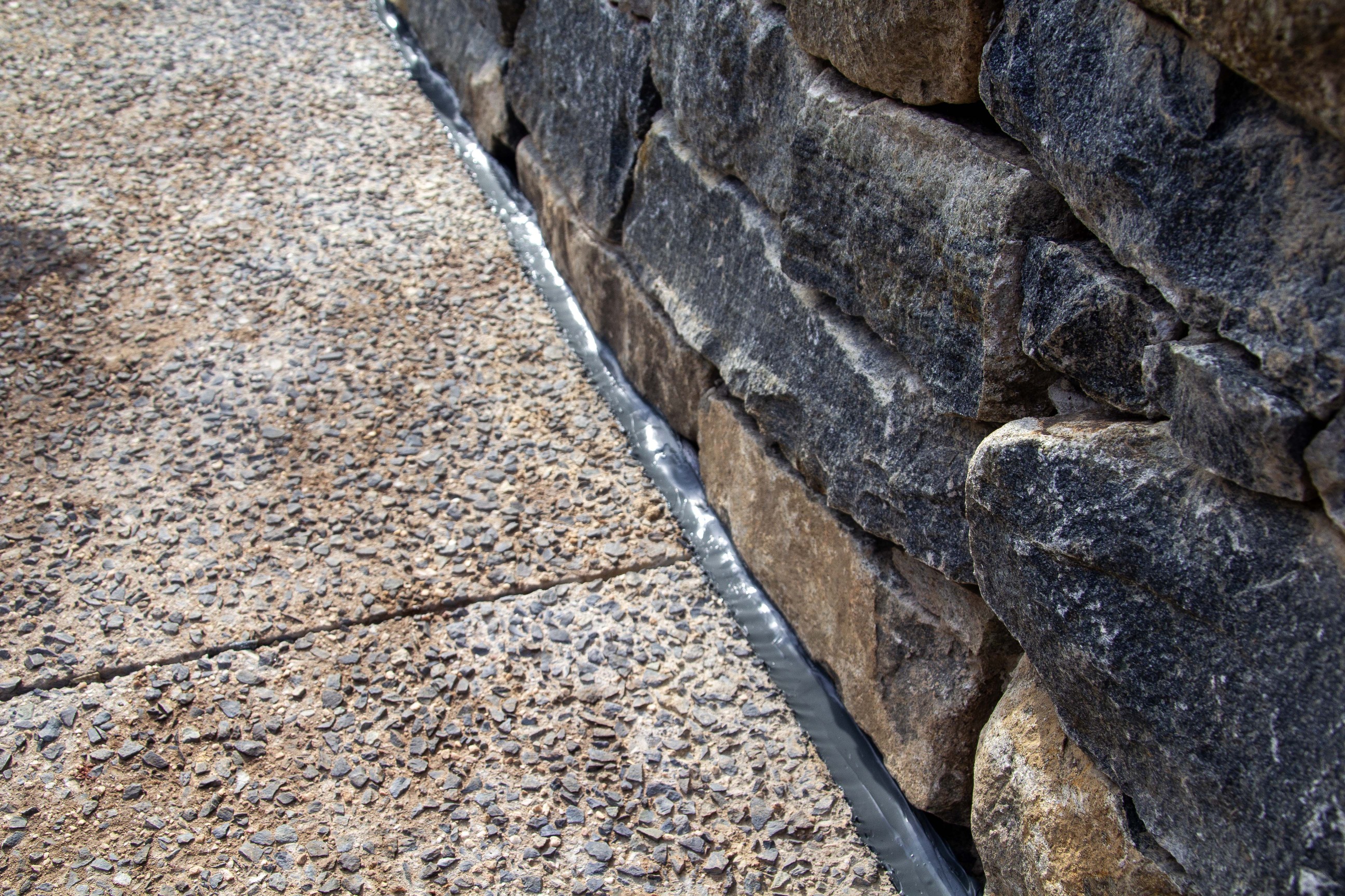 A rock wall and a sidewalk meet diagonally in the middle of the photograph. There is a black-gray line of sealant where the sidewalk meets the rock wall.