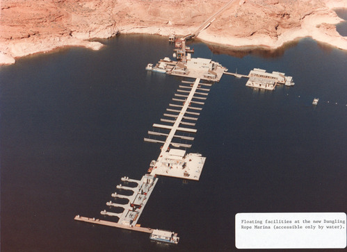 Aerial view of a calm desert lake. A set of boat slips and fuel pumps on a dock are anchored to the back of a sandstone canyon. Label in the Corner: Floating facilities at the new Dangling Rope Marina (accessible only by water).