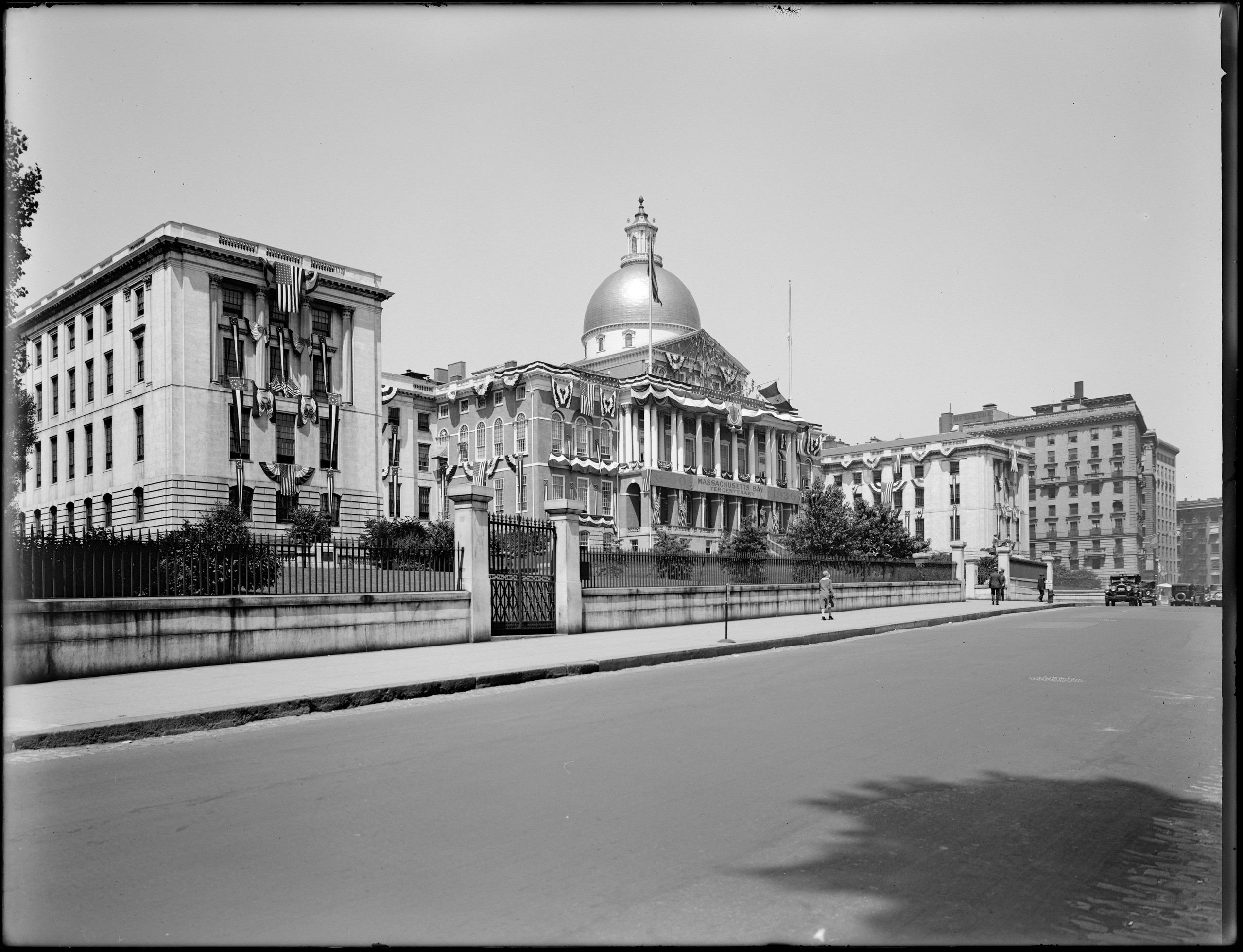 Black and white photograph of the Massachusetts State House from Beacon Street. Building is adorned with decorative flags. A few pedestrians are on the sidewalk, and a small number of cars are on the street.