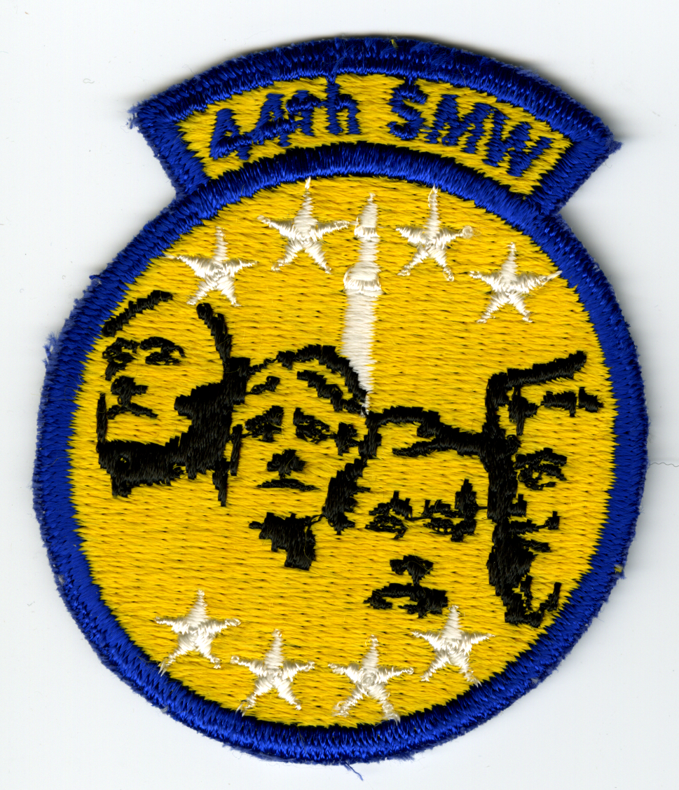 Circular gold with blue trim and tab on top with 44th SMW" in blue on gold. In circle is black outline of heads on Mt. Rushmore with white missile on top center above heads. Four (4) white 5-pointed stars along top and bottom of heads."

