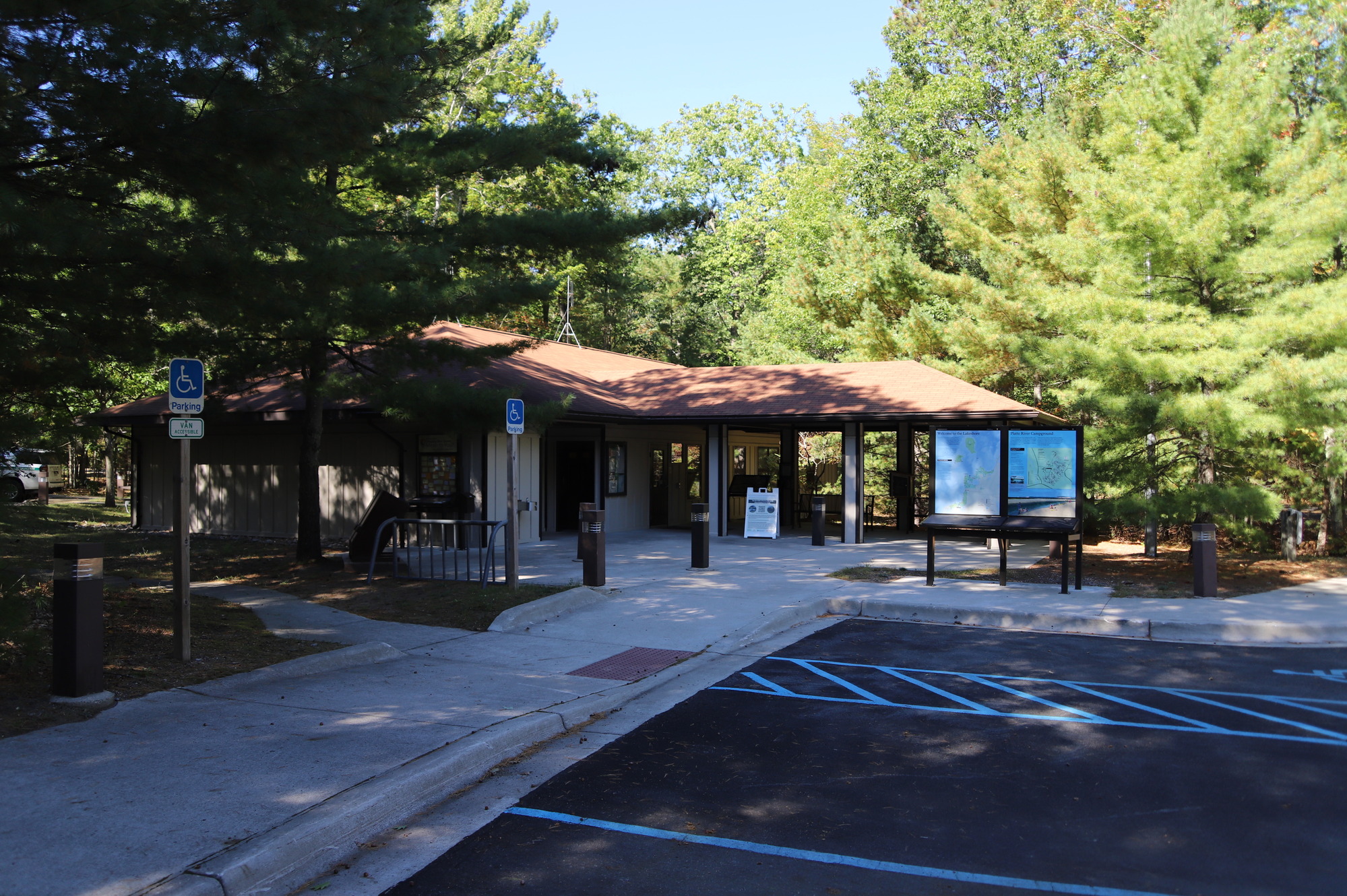 A tan building with a brown shingled roof is surrounded by green trees.  There is a black paved parking area and concrete sidewalk in front of the building.  Visitor Information panels stands in front of the building.  