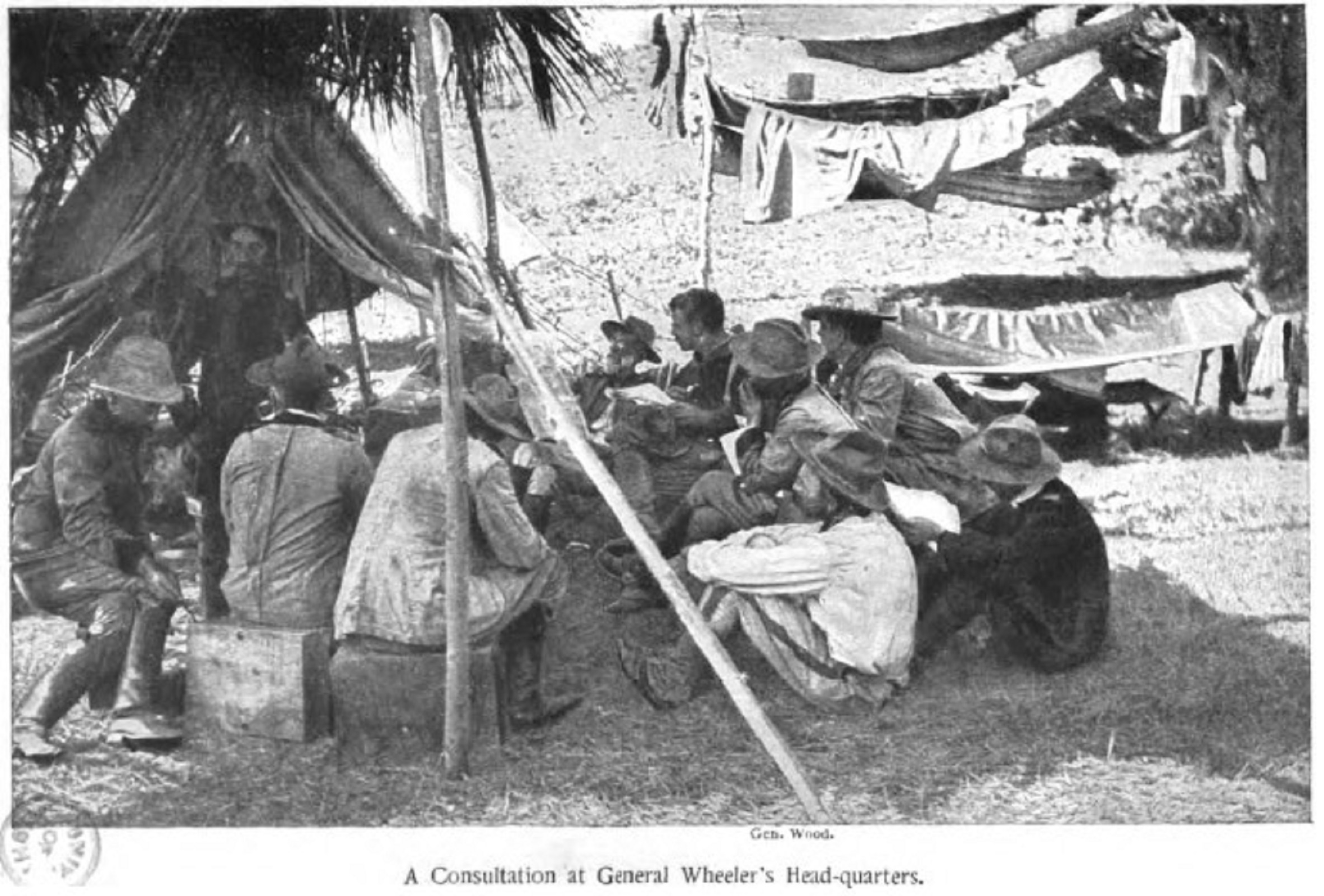Black and white photograph of 10 men gathering in the shade of a tent. some men sit on crates, while others sit on ground. all attention is directed towards General Wheeler. Captioned: A consultation at General Wheeler's Head-quarters. 