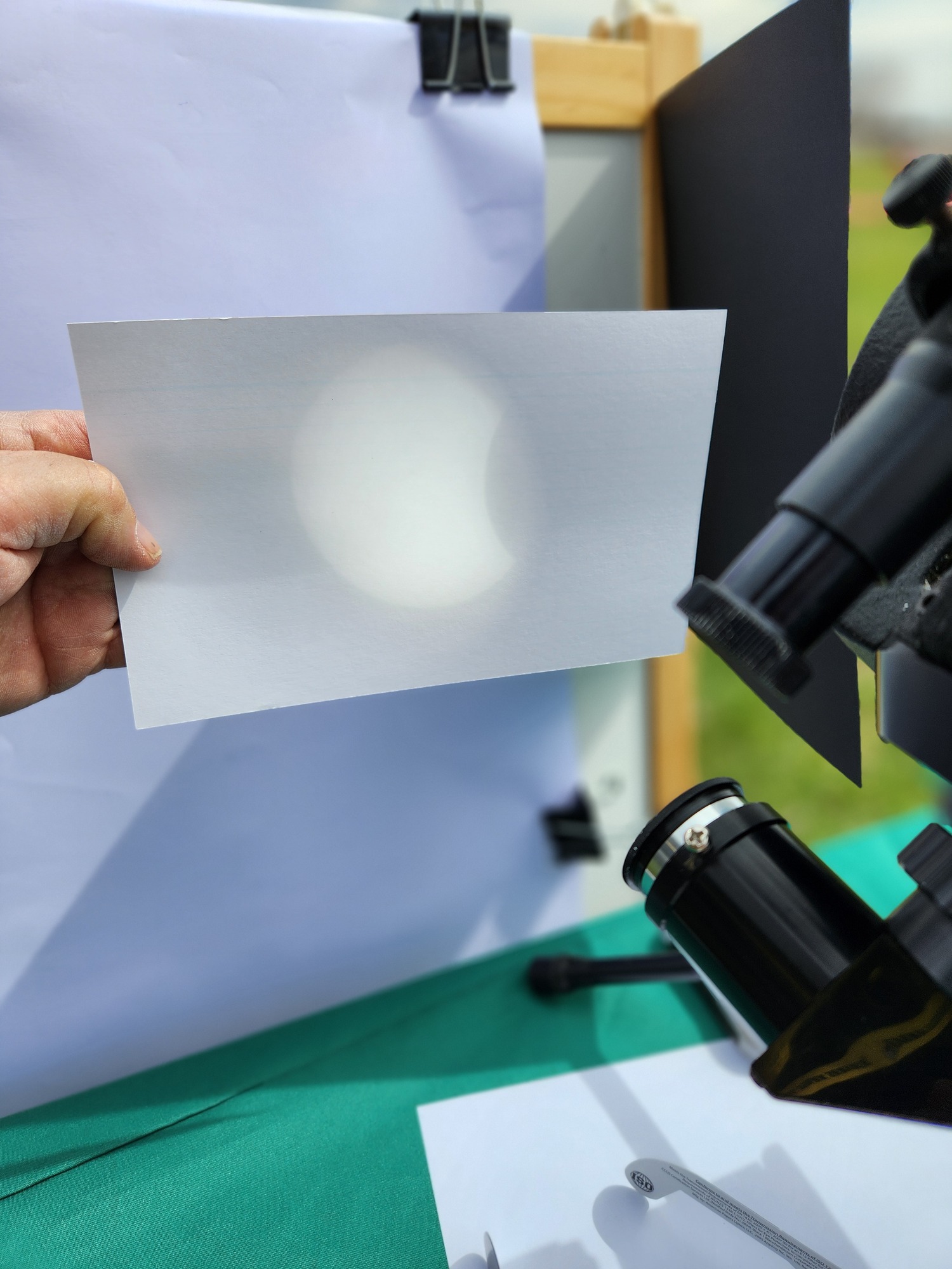 A white sheet of paper held in front of a telescope eyepiece. On it is a bright, nearly full, glowing circle covered by a dark rounded shadow.