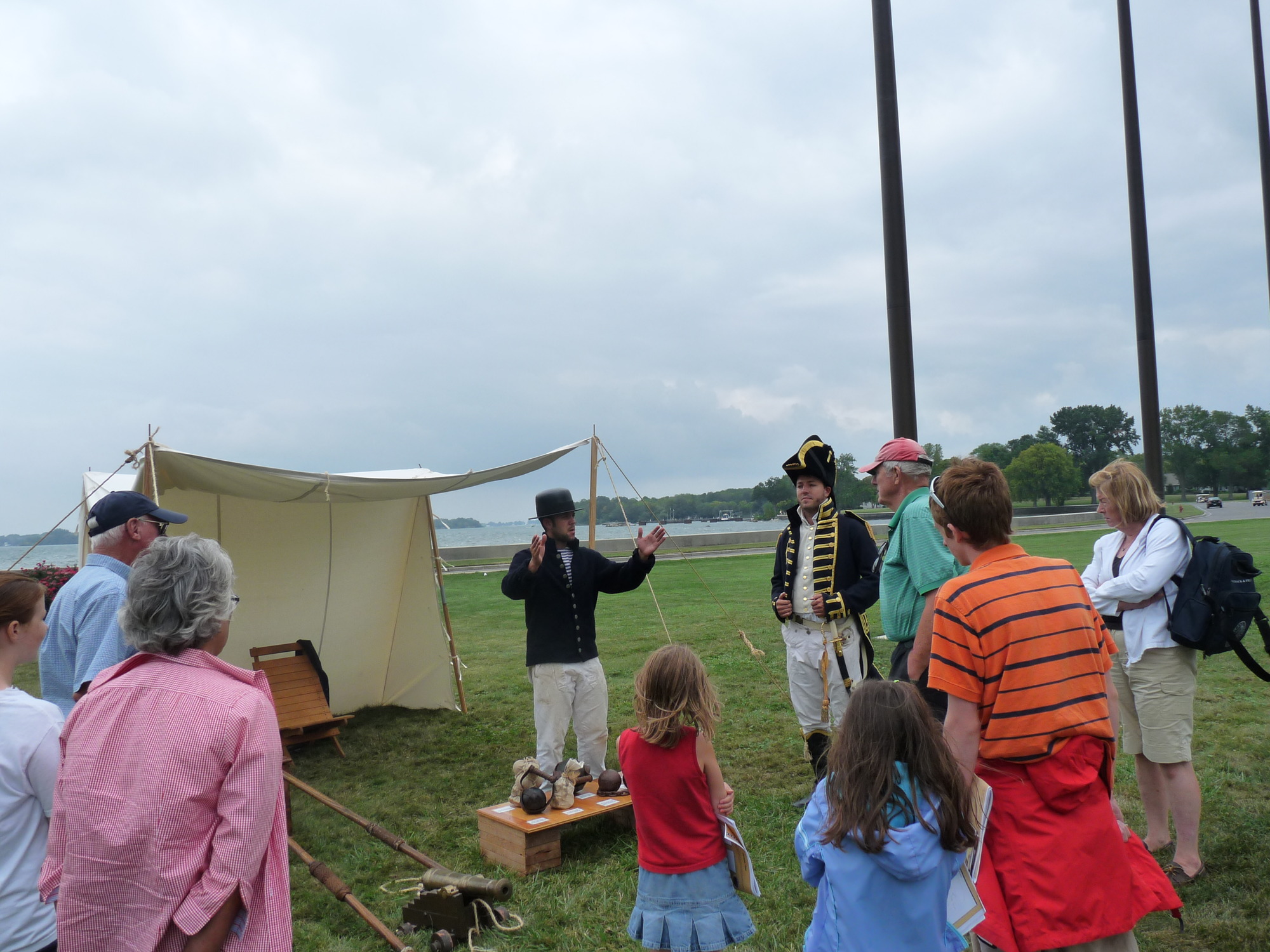 A man dressed as a sailor from the War of 1812,  navy blue coat partially buttoned with white and blue striped shirt, white pants, and a black painted hat,  stands in the center of the picture with hands out front as if holding something 24 inches long between them. At his feet is a small table with various shot fired from naval artillery. He is surrounded by a group of people ranging in age young to old.