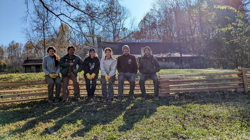 A group of six young people smiling and posing for a photo, sit on a newly constructed fence with blue skies above them. Trees, sign for the Nancy Lincoln Inn and the wooden Inn structure are visible in the background.