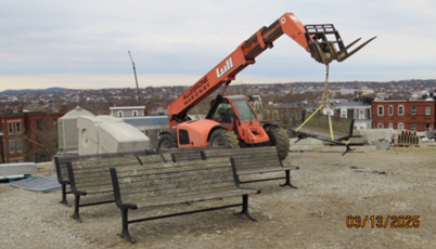 A forklift lifts a wooden bench in an open construction site. Other wooden benches line the dirt area. 