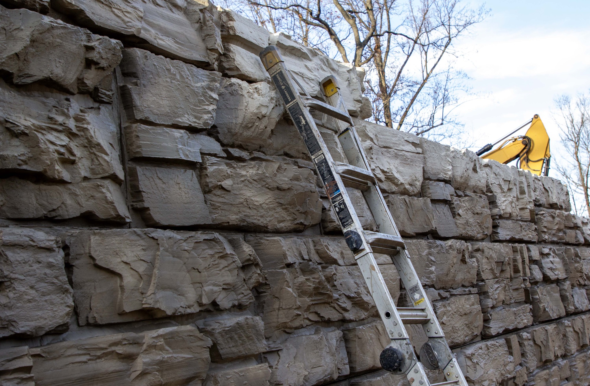 : A large stone rock wall with a ladder in the middle of the image. The top of a construction vehicle appears in the upper right of the photograph. 