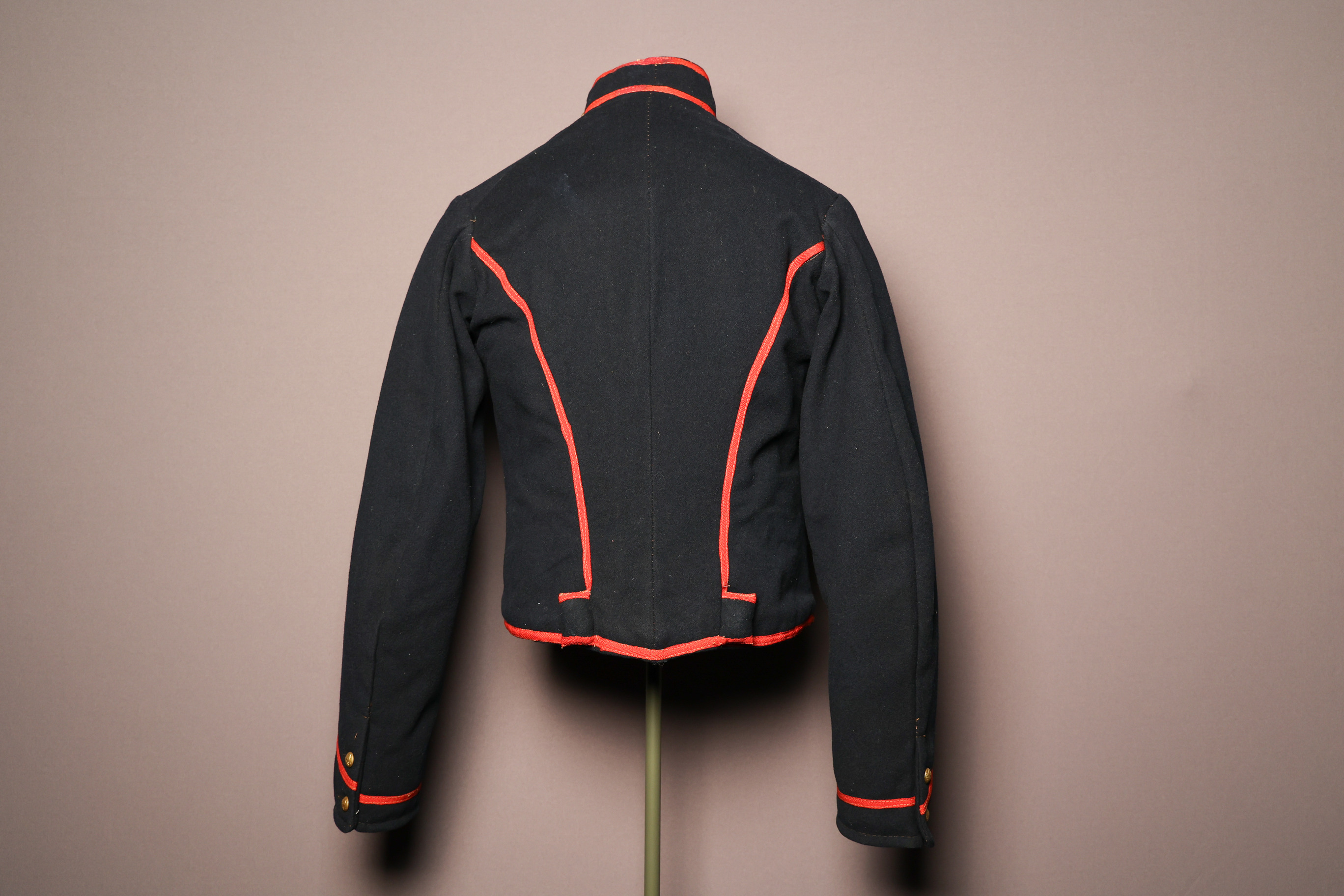 Color image of the back of a field artilleryman Civil War uniform shell jacket. Jacket is navy blue with orange piping on the collar, sleeve cuff, back side seams, and bottom hem. Two small gold metal, circular buttons accent each of the sleeve cuffs. Jacket is a woolen texture.